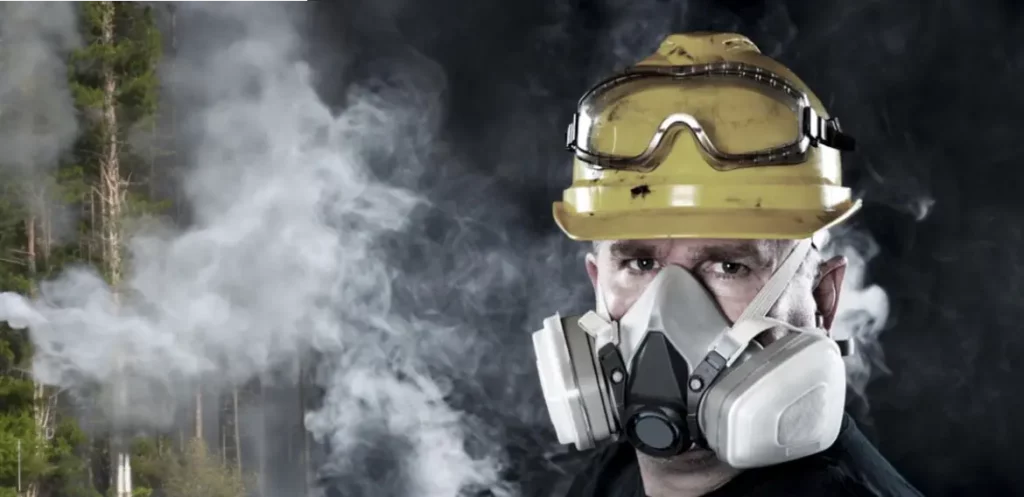 Man in a respirator mask wearing a hard hat with smoky trees in the background